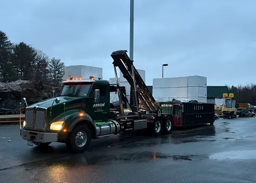 Arrow Container Services truck with hook lift mechanism delivering a waste dumpster in a commercial setting, surrounded by stacked materials and equipment.